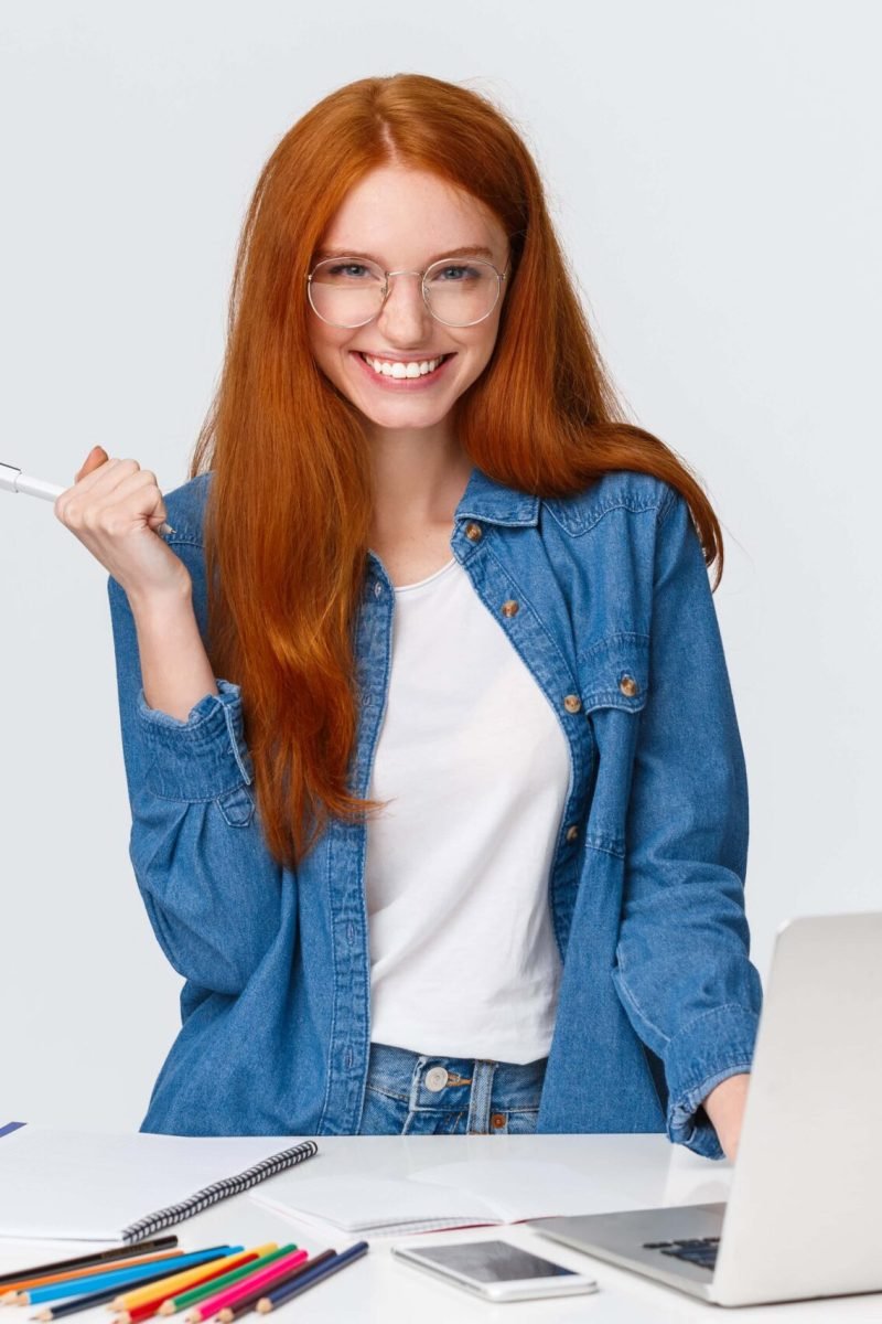 sassy-confident-good-looking-charismatic-redhead-female-near-laptop-table-with-drawing-supplies-smiling-cheeky-mystic-camera-standing-white-background-have-excellent-plan (1)