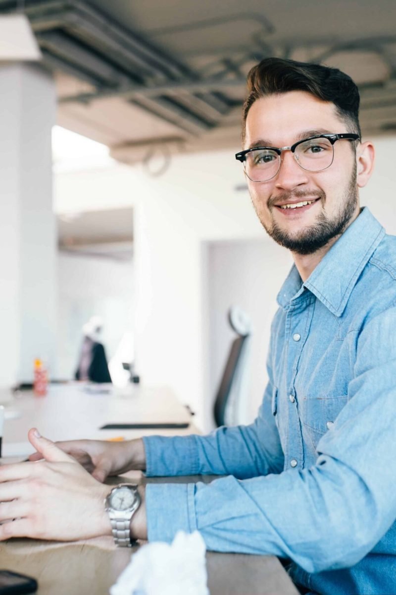 young-dark-haired-man-glasses-is-working-with-computer-his-desktop-office-he-wears-blue-shirt-smiles-camera-view-from-side (1)